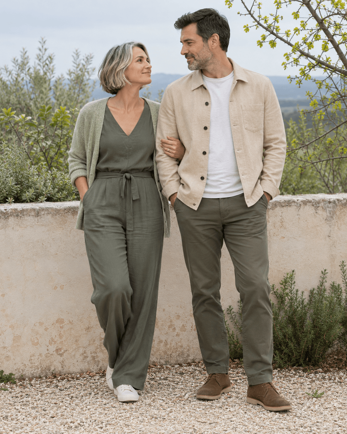 AI-generated concept: two fictional adults beside a low pale garden wall, sparse fresh leaves beyond, one sand light jacket and one muted green outfit, overcast daylight, full-length, no dense blossoms.