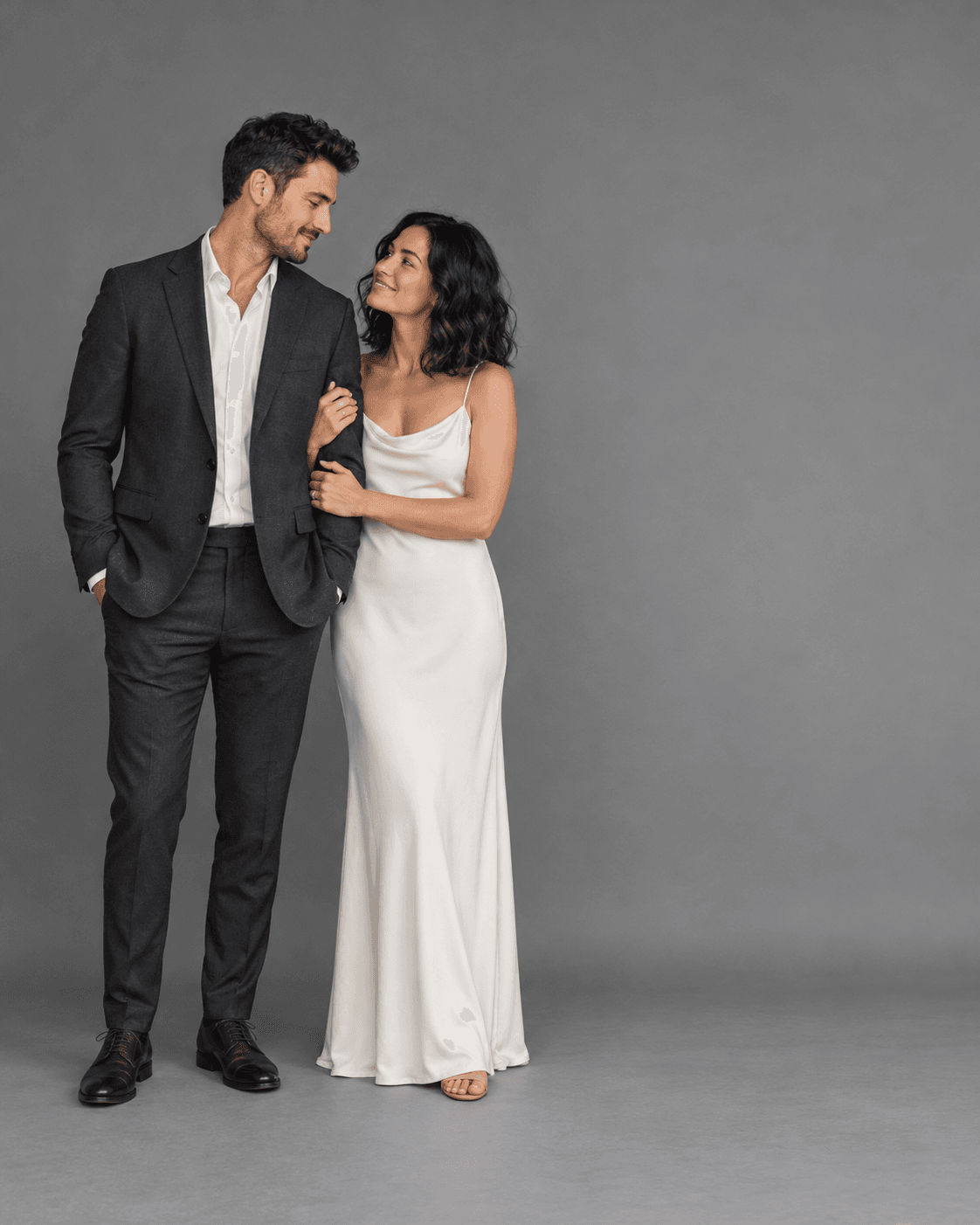 AI-generated concept: two fictional adults against a medium-gray studio backdrop, full-length restrained pose, generous negative space on one side, charcoal and ivory garments.