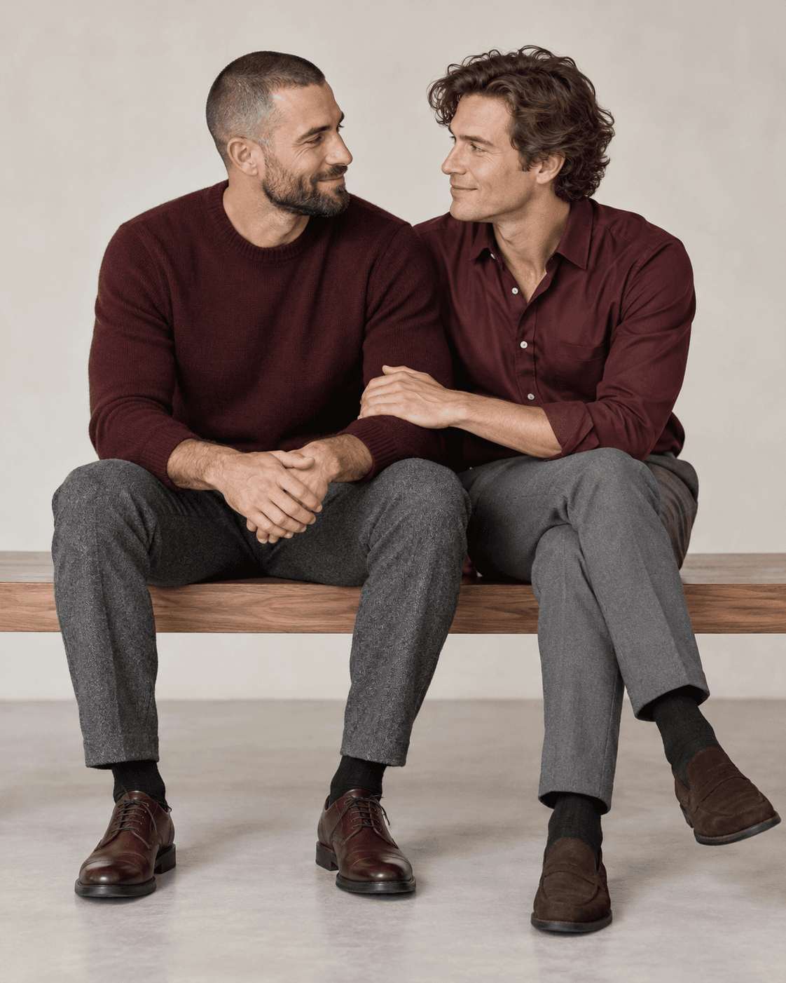 AI-generated concept: two fictional adults seated on a broad simple bench against a pale studio background, intentional upright postures and small inward turns, refined burgundy and gray separates, full seated view.