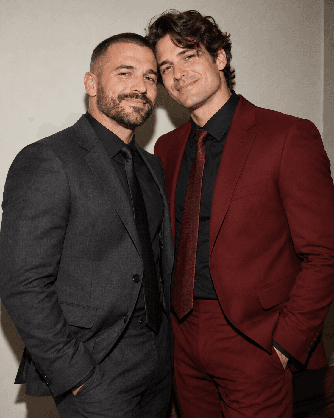 AI-generated concept: two fictional adults in formal charcoal and deep red clothing, relaxed shared smile, direct-flash-inspired lighting, simple indoor wall, three-quarter crop.