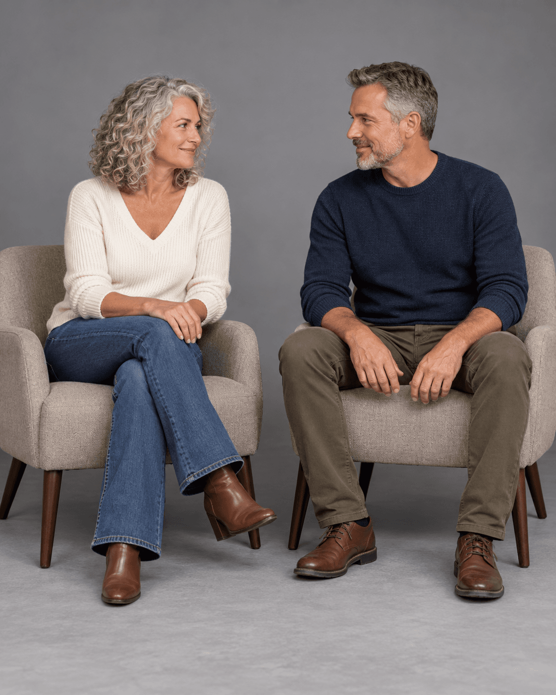 AI-generated concept of two fictional adults seated on separate broad chairs angled slightly inward against a gray studio backdrop, sharing a glance, with one adult crossing their legs.